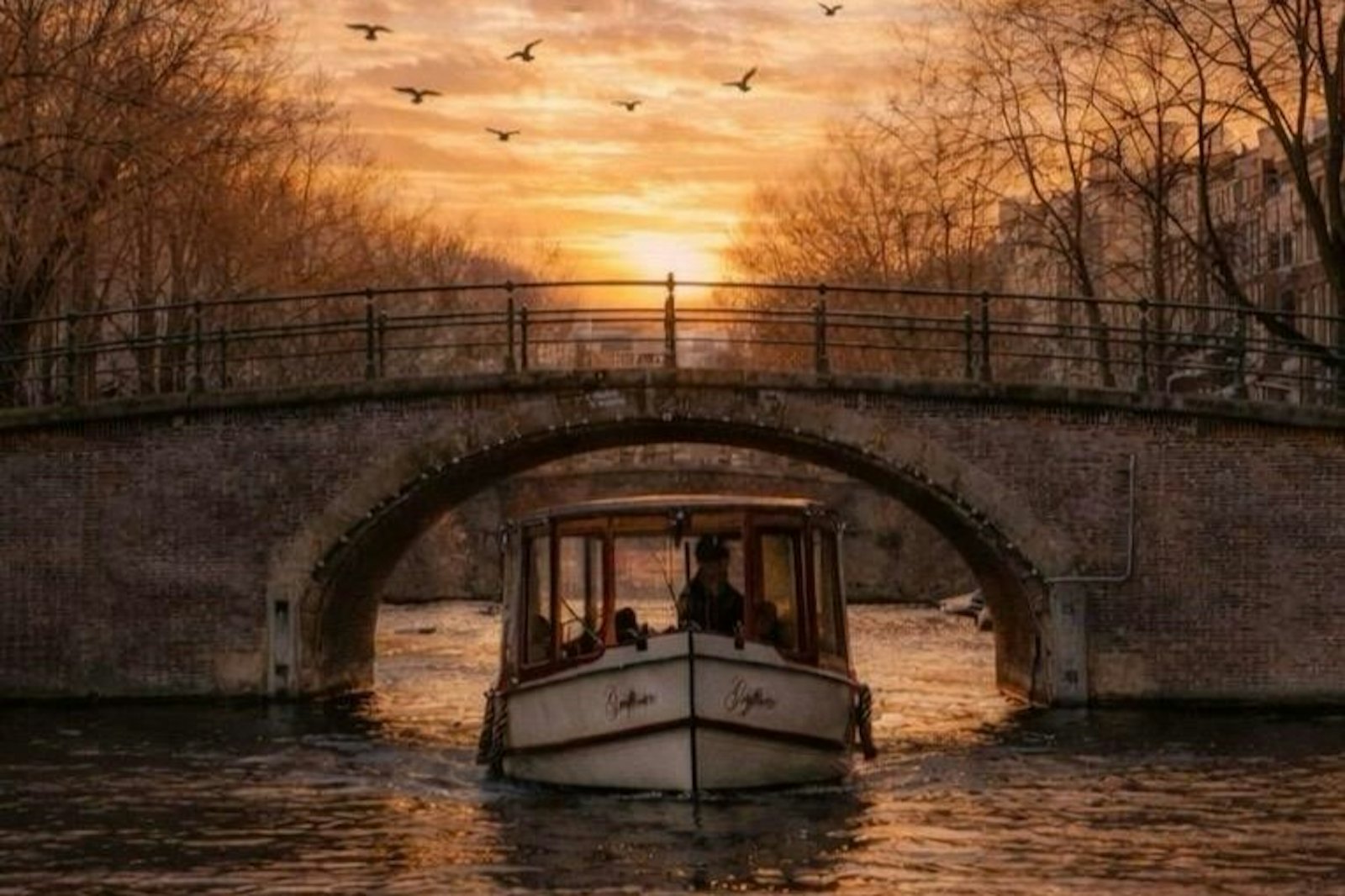 Open canal boat passing under a stone bridge on an Amsterdam canal at sunset