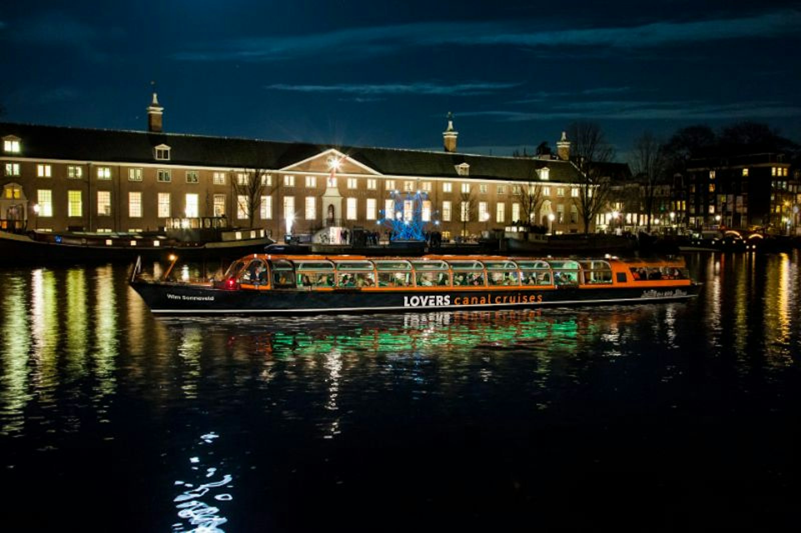 Lit saloon canal boat passing the H’ART Museum on the Amstel during an evening canal cruise